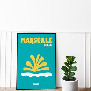 May include: A framed poster with a teal background features the words "MARSEILLE BELLE" in yellow, with a stylized yellow design and the word "FRANCE" at the bottom. A small potted plant sits to the right.