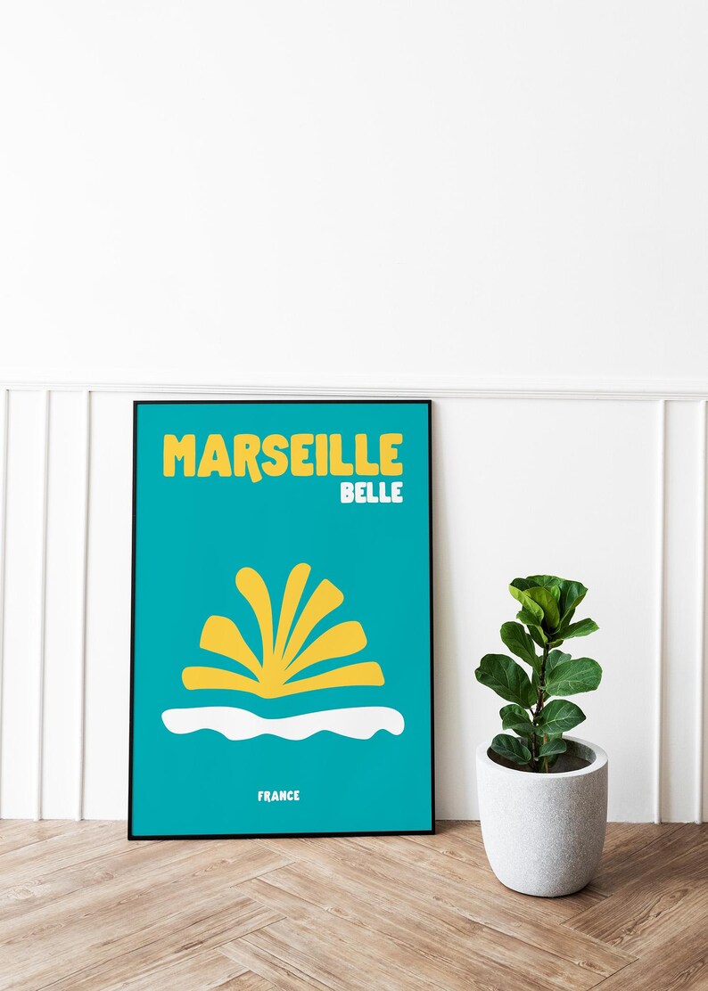 May include: A framed poster with a teal background features the words "MARSEILLE BELLE" in yellow, with a stylized yellow design and the word "FRANCE" at the bottom. A small potted plant sits to the right.