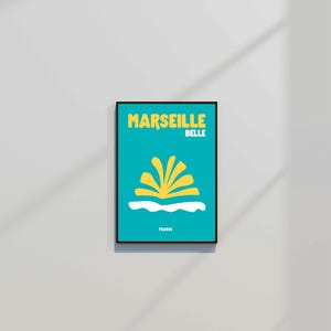 May include: A framed art print with a teal background features the words "MARSEILLE BELLE" in yellow, above a yellow and white graphic. The word "FRANCE" is printed at the bottom. The print is in a black frame.
