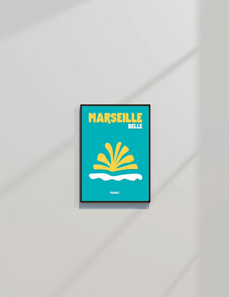May include: A framed art print with a teal background features the words "MARSEILLE BELLE" in yellow, above a yellow and white graphic. The word "FRANCE" is printed at the bottom. The print is in a black frame.