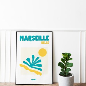 May include: A framed art print with the words "MARSEILLE BELLE" in teal and yellow. The print features a yellow sun, teal and yellow abstract shapes, and the word "FRANCE". A potted plant sits to the right.
