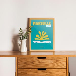 May include: A framed poster with the words "MARSEILLE BELLE" in yellow on a teal background. The poster features a yellow and white graphic design and the word "FRANCE" at the bottom. A white pitcher with greenery sits to the left.