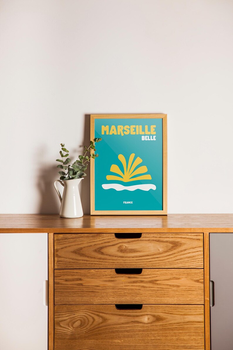 May include: A framed poster with the words "MARSEILLE BELLE" in yellow on a teal background. The poster features a yellow and white graphic design and the word "FRANCE" at the bottom. A white pitcher with greenery sits to the left.