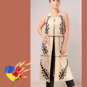 May include: A long, sleeveless beige vest with black embroidered floral patterns. The vest has a vertical design with decorative stitching and a tie closure. The model is wearing black trousers. The background is a solid brown colour.
