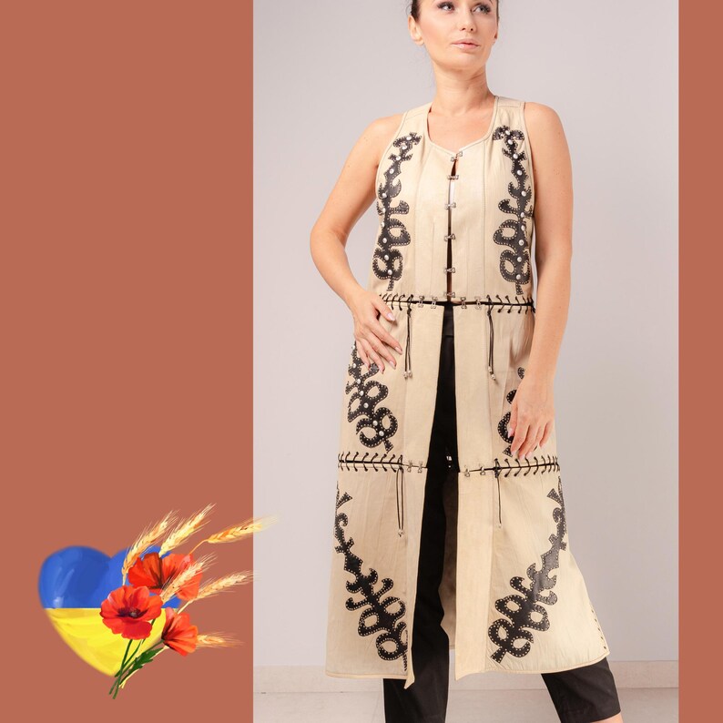 May include: A long, sleeveless beige vest with black embroidered floral patterns. The vest has a vertical design with decorative stitching and a tie closure. The model is wearing black trousers. The background is a solid brown colour.