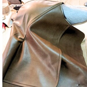 May include: Close-up of a brown leather garment, possibly a jacket or coat, showing detailed stitching and a smooth, textured surface. The image highlights the craftsmanship and material quality of the product.