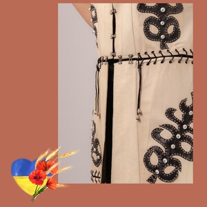 May include: Cream-coloured sleeveless garment with black leather appliqu&eacute; and silver-tone hardware. The design features decorative black leather shapes with silver studs and a black belt. The garment has a vertical black stripe down the front.