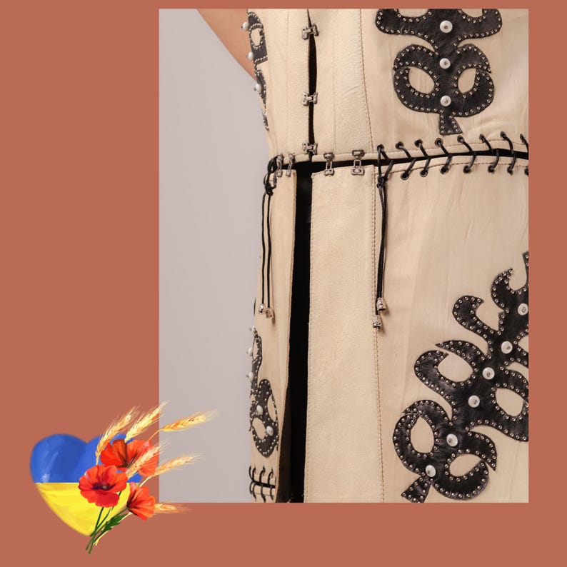 May include: Cream-coloured sleeveless garment with black leather appliqu&eacute; and silver-tone hardware. The design features decorative black leather shapes with silver studs and a black belt. The garment has a vertical black stripe down the front.