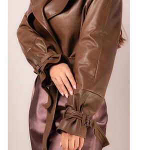 May include: A brown leather trench coat with a belted waist and cuffs. The coat has a classic design with a lapel collar and long sleeves. The cuffs are detailed with buckles and straps. The coat is made of a smooth, supple leather.