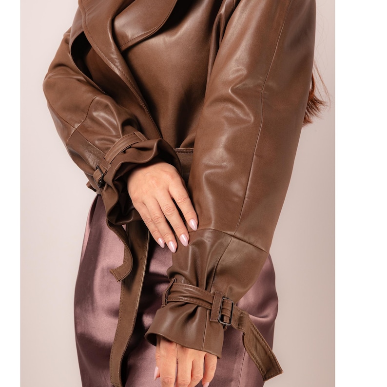 May include: A brown leather trench coat with a belted waist and cuffs. The coat has a classic design with a lapel collar and long sleeves. The cuffs are detailed with buckles and straps. The coat is made of a smooth, supple leather.