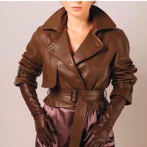 May include: A brown leather jacket with a wide collar, belted waist, and long sleeves. The jacket has a classic trench coat design. The model is also wearing matching brown leather gloves.