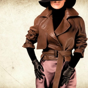 May include: A brown leather jacket with a wide collar and belt, paired with a black turtleneck, gloves, and a wide-brimmed hat. The outfit includes a satin skirt. The jacket has a tailored fit.