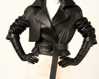May include: A black leather jacket with a belted waist and long sleeves. The jacket features a wide collar and is worn with matching long gloves. The outfit is completed with a cream-coloured skirt.