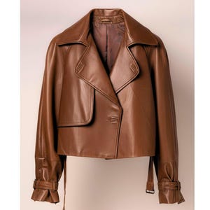 May include: A brown leather jacket with a wide collar, button closure, and long sleeves. The jacket features a flap pocket and adjustable cuffs with buckle details. The overall design is a classic, tailored style.