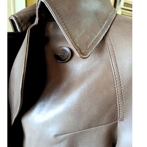 May include: Close-up of a brown leather trench coat. The coat features a notched collar, a large button, and visible stitching details. The leather has a smooth texture and a warm, neutral tone.