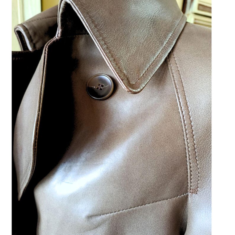 May include: Close-up of a brown leather trench coat. The coat features a notched collar, a large button, and visible stitching details. The leather has a smooth texture and a warm, neutral tone.