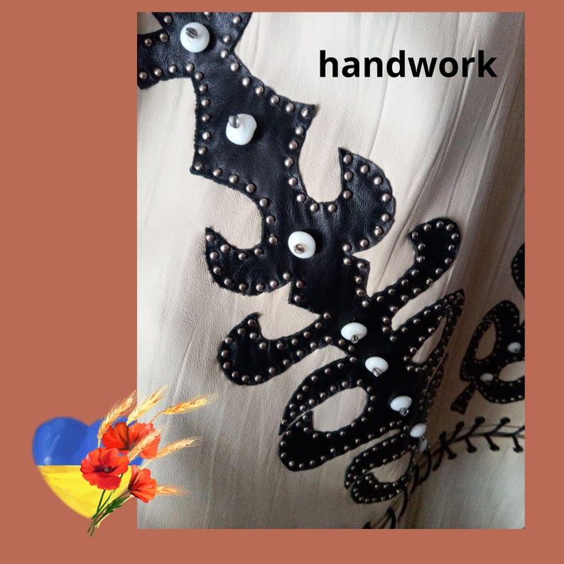 May include: Close-up of a beige fabric with black leather applique. The applique is adorned with silver studs and white pearl-like accents. The word "handwork" is printed on the fabric.