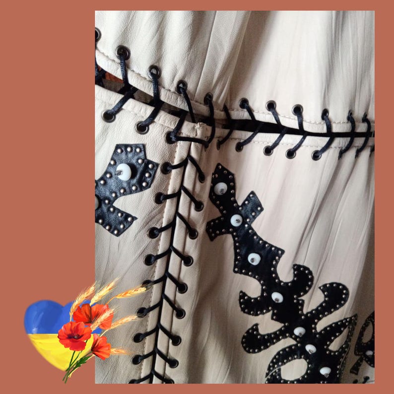 May include: Close-up of a beige leather garment with black decorative stitching and embellishments. The design includes black leather cutouts with silver studs and white accents. The garment has a unique, handcrafted appearance.