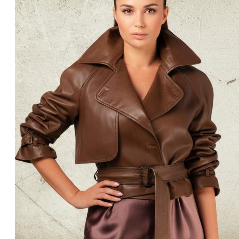 May include: A brown leather jacket with a wide collar, belted waist, and short sleeves. The jacket has a cropped design and is worn over a satin dress. The overall style is classic and sophisticated.