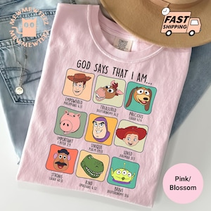 May include: Pink Comfort Colors t-shirt with a graphic featuring cartoon characters and the text "GOD SAYS THAT I AM...". Each character is labeled with a positive adjective and a Bible verse reference. The shirt is folded on a pair of blue jeans.