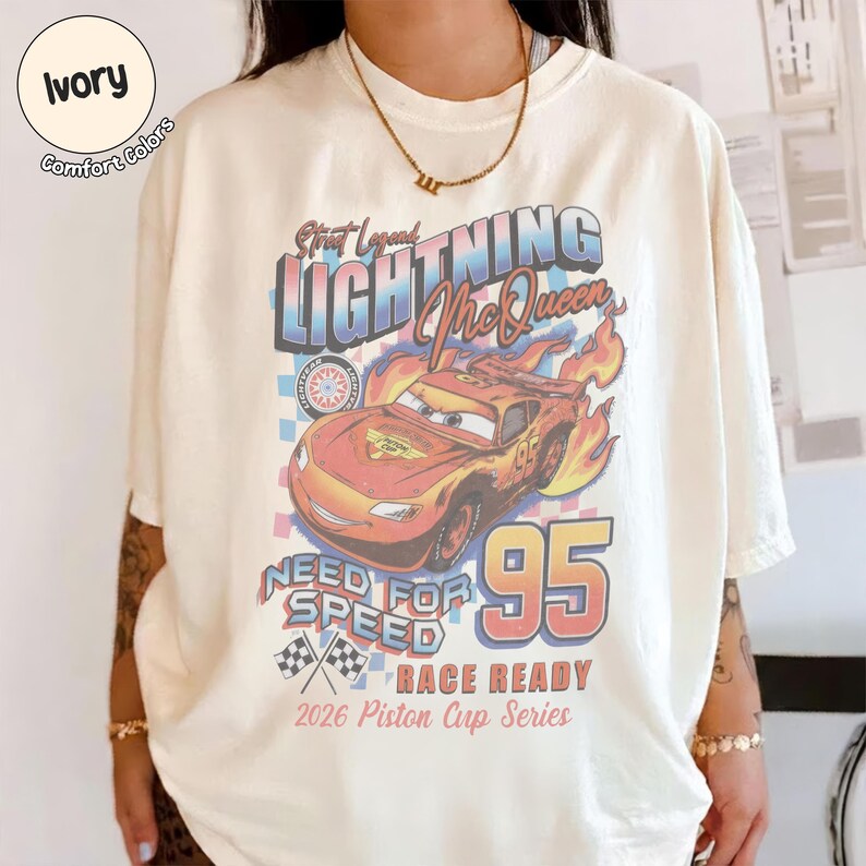 May include: Ivory-colored t-shirt featuring a graphic of Lightning McQueen from the movie Cars. The design includes the text "Street Legend LIGHTNING McQueen 95 NEED FOR SPEED RACE READY 2026 Piston Cup Series". The shirt has short sleeves and a crew neck.