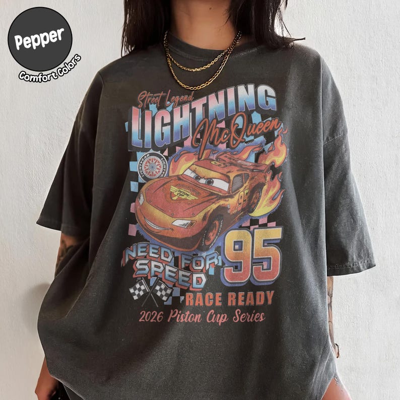 May include: Dark gray t-shirt featuring a graphic of Lightning McQueen from the movie Cars. The design includes the text "Street Legend Lightning McQueen 95 Need for Speed Race Ready 2026 Piston Cup Series". The shirt is a Comfort Colors brand.