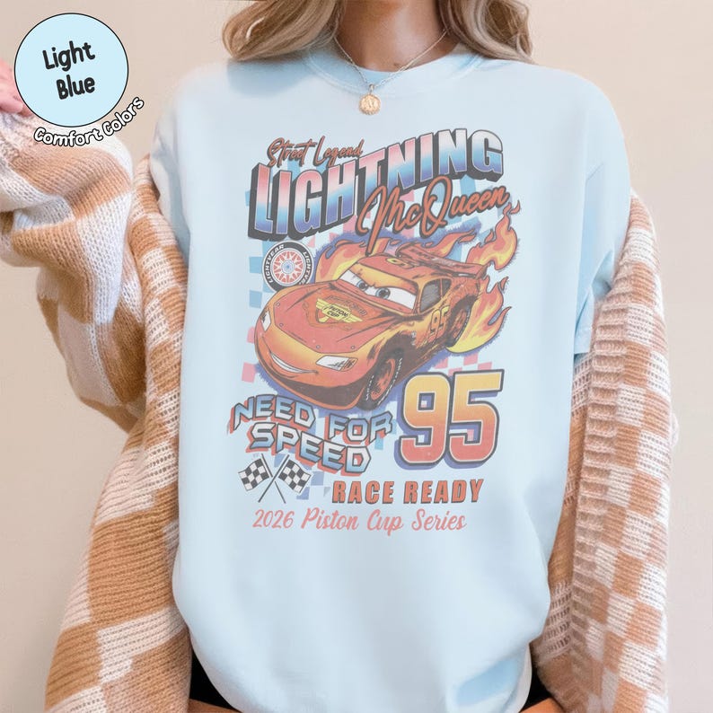 May include: Light blue sweatshirt featuring a graphic of Lightning McQueen from the movie Cars. The design includes the text "Street Legend Lightning McQueen", "Need for Speed", and the number "95". The sweatshirt is labeled "Race Ready" and "2026 Piston Cup Series".