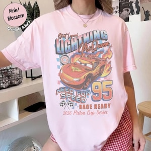 May include: Pink t-shirt featuring a graphic of Lightning McQueen from the movie Cars. The design includes the text "Lightning McQueen", "Need for Speed", and "95". The shirt also has the text "Race Ready" and "2026 Piston Cup Series".