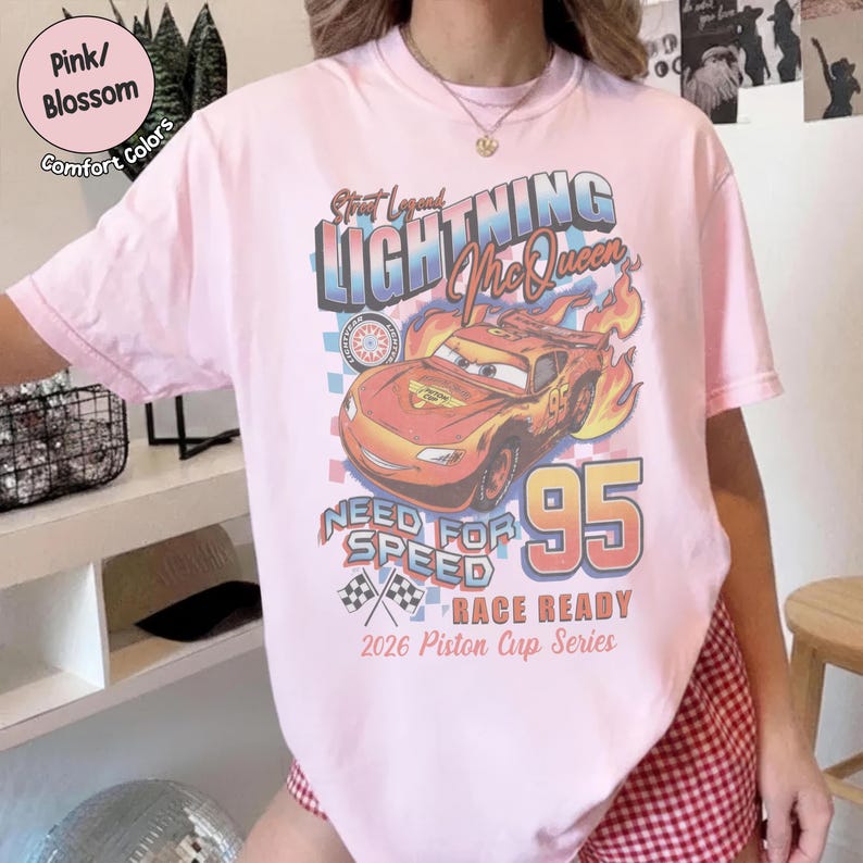 May include: Pink t-shirt featuring a graphic of Lightning McQueen from the movie Cars. The design includes the text "Lightning McQueen", "Need for Speed", and "95". The shirt also has the text "Race Ready" and "2026 Piston Cup Series".