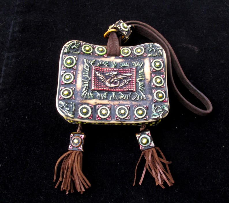 May include: A small, rectangular, decorative pouch with a brown leather strap. The pouch is decorated with green, red, and gold accents and a dragon design. The pouch has two brown tassels.