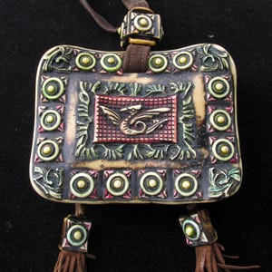 May include: A white ceramic pendant with a dragon design, surrounded by green and gold accents. The pendant is attached to a brown leather cord with gold beads.