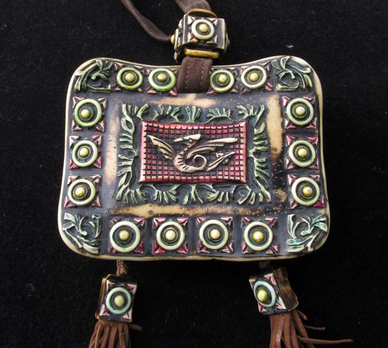 May include: A white ceramic pendant with a dragon design, surrounded by green and gold accents. The pendant is attached to a brown leather cord with gold beads.