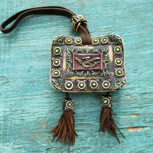 May include: A small, rectangular, cream-colored pouch with a brown leather strap and tassels. The pouch has a raised, ornate design with a dragon in the center and a border of green and gold circles.