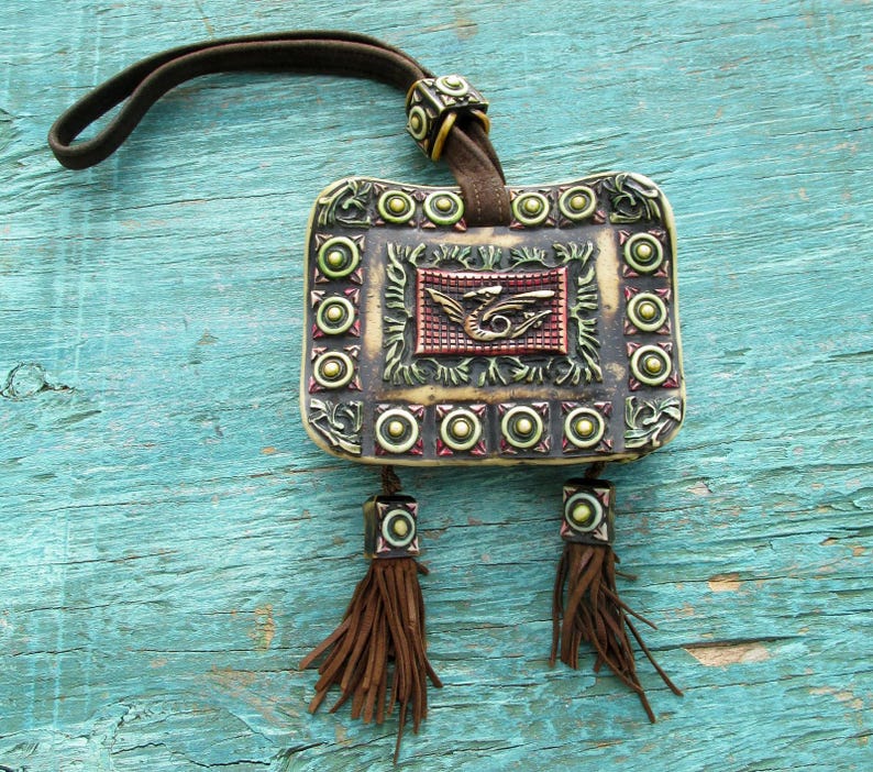May include: A small, rectangular, cream-colored pouch with a brown leather strap and tassels. The pouch has a raised, ornate design with a dragon in the center and a border of green and gold circles.