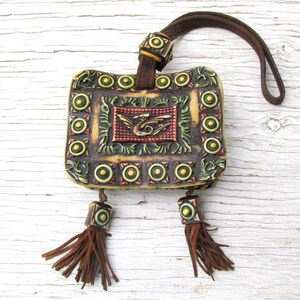 May include: A small, square, leather pouch with a brown strap and tassels. The pouch is decorated with a dragon design and gold and green accents.