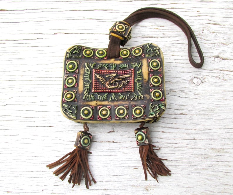 May include: A small, square, leather pouch with a brown strap and tassels. The pouch is decorated with a dragon design and gold and green accents.