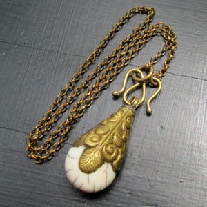 May include: A vintage-style necklace with a brass-colored chain and pendant. The pendant features a teardrop shape with a white base and ornate gold-colored metal detailing. The chain is approximately 24 inches long.