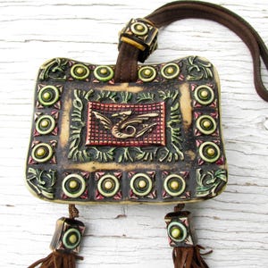 May include: A beige ceramic pouch with a decorative design featuring a dragon, green leaves, and gold circles. The pouch has a brown leather strap and tassels.