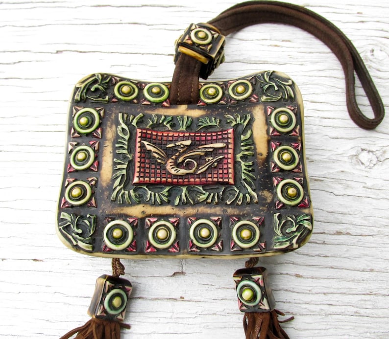 May include: A beige ceramic pouch with a decorative design featuring a dragon, green leaves, and gold circles. The pouch has a brown leather strap and tassels.