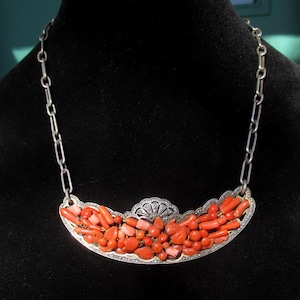 May include: A silver necklace featuring a curved pendant filled with coral-colored beads and stones. The pendant is outlined with a silver filigree design and connected to a silver chain.