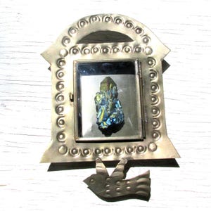 May include: A small, antique-style metal frame with a decorative bird attached. The frame holds a colorful, iridescent crystal. The frame is silver-toned with a decorative pattern around the edges. The crystal has shades of blue, green, and gold.