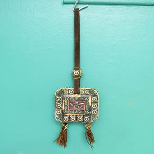 May include: A small, square, decorative pouch with a green, brown, and yellow pattern. The pouch has a brown leather strap with tassels and is hanging from a hook.