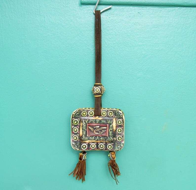 May include: A small, square, decorative pouch with a green, brown, and yellow pattern. The pouch has a brown leather strap with tassels and is hanging from a hook.