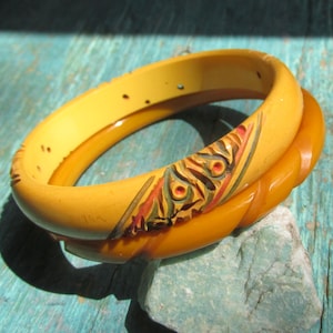 May include: Two stacked bangle bracelets in shades of yellow and amber. The top bangle is a smooth, pale yellow with small dark spots and a carved, painted floral-like design. The bottom bangle is a deeper, translucent amber with a twisted texture.