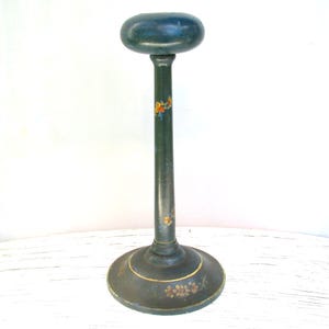 May include: A dark green hat stand with a round base and a tall, slender stem. The top is a rounded oval shape. The stand is decorated with small floral accents in orange, yellow, and red. The base has a gold trim.