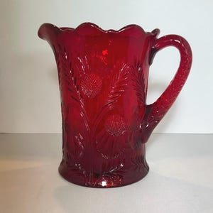 May include: A ruby red glass pitcher featuring a raised, textured thistle and leaf pattern across its exterior. The vessel has a scalloped rim, a curved handle, and a pouring spout, showcasing a translucent finish that catches the light.