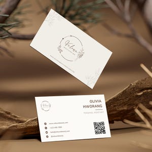 Könnte beinhalten: Zwei Visitenkarten mit minimalistischem Design. Die Karten sind weiß und tragen den Namen "Veluna" in einem kreisförmigen Logo mit floralen Akzenten. Die untere Karte zeigt Kontaktinformationen, einschließlich eines QR-Codes und den Namen "Olivia Hworang, Persönliche Assistentin."