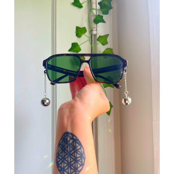 May include: Sunglasses with a dark tortoise shell frame and green lenses. Each side of the sunglasses has a silver chain with a small disco ball charm attached. The sunglasses are held up by a hand.