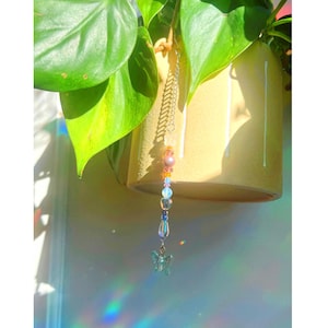 Puede incluir: Un colgador de plantas decorativo con una cadena de cuentas y un pequeño dije de mariposa transparente. La cadena presenta cuentas coloridas en tonos rosa, naranja, azul y transparente. El colgador de plantas cuelga frente a una maceta amarilla con hojas verdes.
