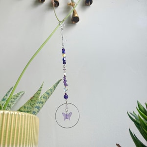 Puede incluir: Un atrapasoles decorativo con un marco circular transparente y un dije de mariposa morada. El atrapasoles está suspendido por una cadena plateada y adornado con cuentas moradas, azules y transparentes. El fondo incluye plantas y una pared.